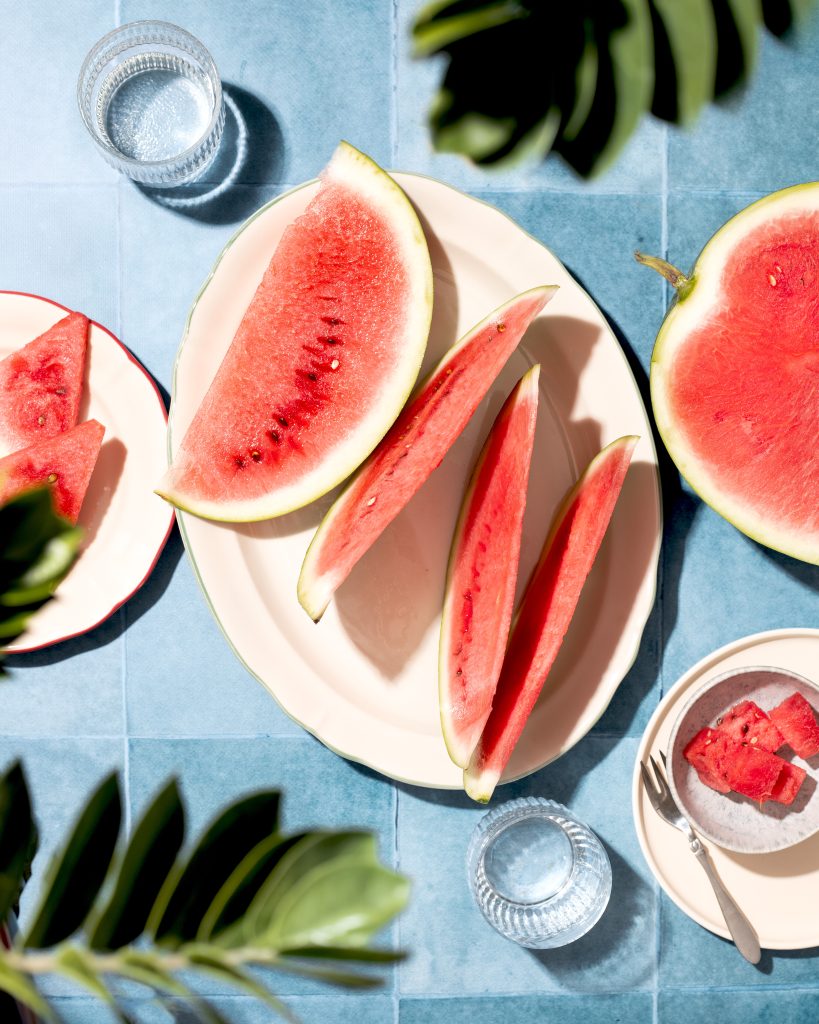 Editorial flat lay photograph of fresh watermelon slices arranged on white plates over a blue tiled background. Tropical leaves and bright natural sunlight create a colorful summer food scene with a clean editorial aesthetic.