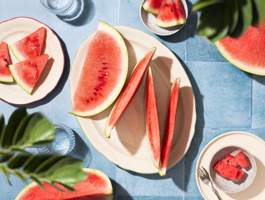 Editorial flat lay photograph of fresh watermelon slices arranged on white plates over a blue tiled background. Tropical leaves and bright natural sunlight create a colorful summer food scene with a clean editorial aesthetic.