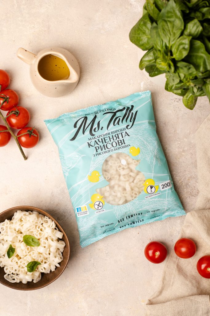 flat lay of a package of rice pasta on a beige background, next to a bowl of cooked pasta, tomatoes and basil