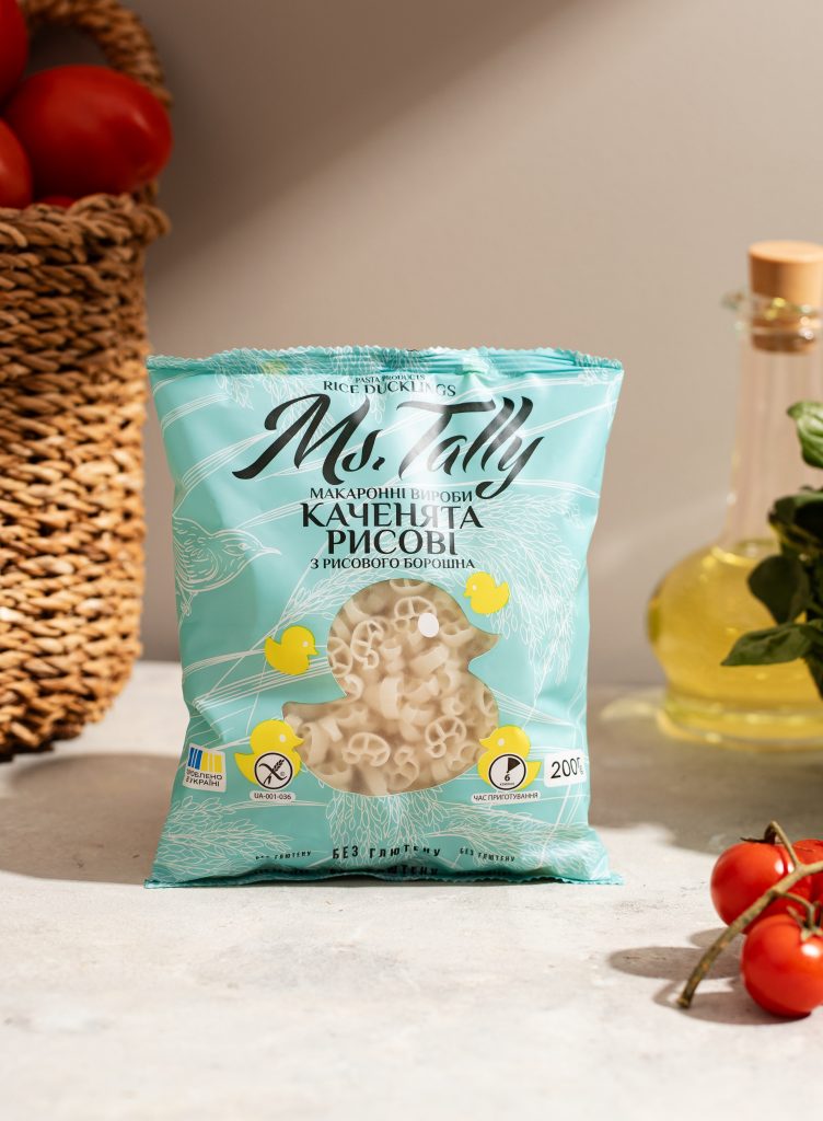 a package of rice baby pasta, behind it on the left is a basket of tomatoes, and on the right is a bottle of oil